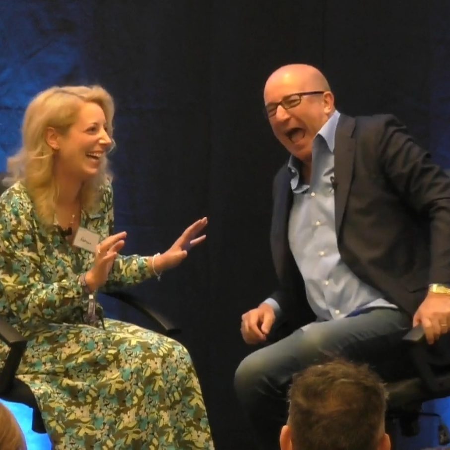 Paul McKenna and Rebecca Rebecca, in a patterned dress, and Paul McKenna in a blazer engage in conversation on stage.