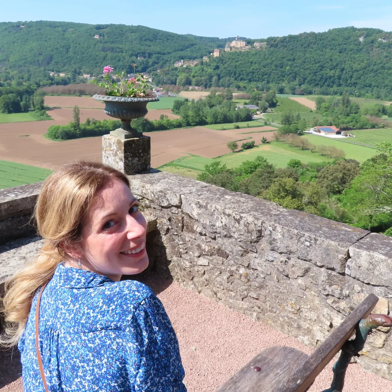 Your Hypnotherapist, Rebecca A smiling woman sits on a stone terrace overlooking lush green fields and hills.