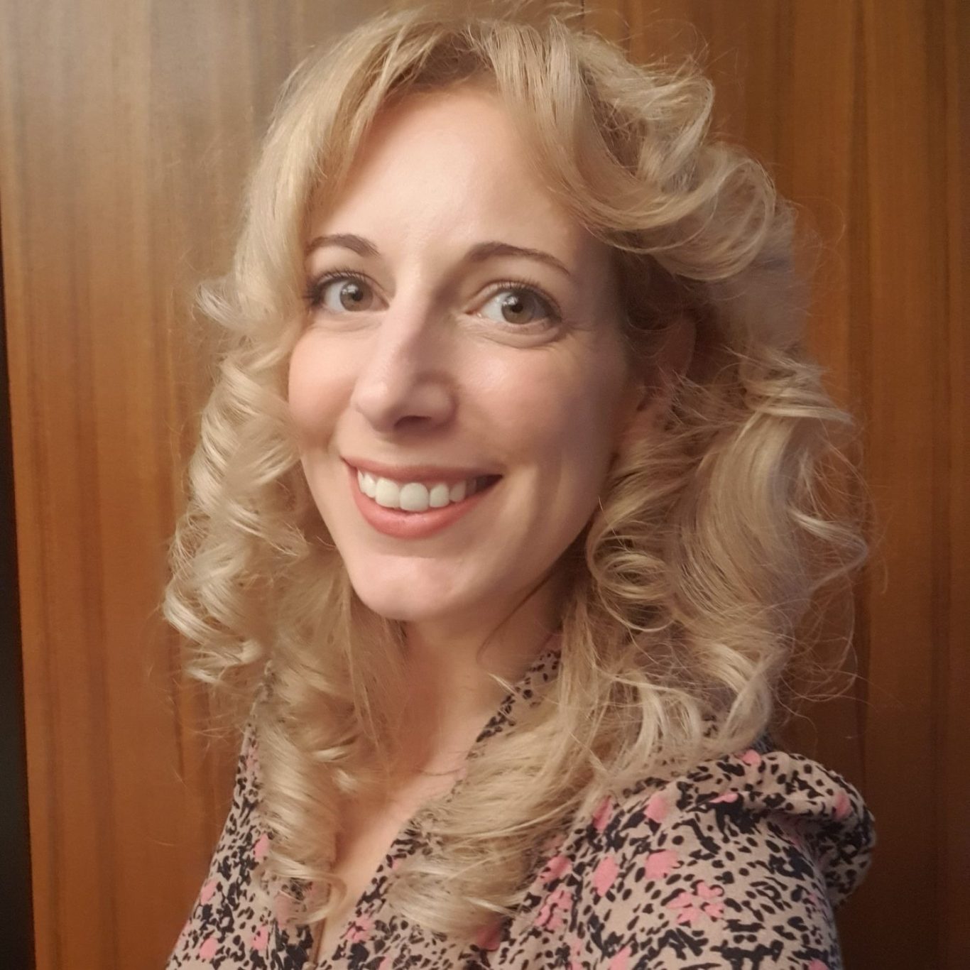 Your Hypnotherapist, Rebecca Rebecca, with curly blonde hair smiling, wearing a floral dress against a wooden background.